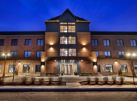 A large, modern building with a dark facade and a central entrance is illuminated by exterior lights at dusk.
