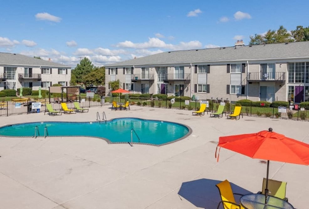 A swimming pool surrounded by chairs and umbrellas in front of apartment buildings.