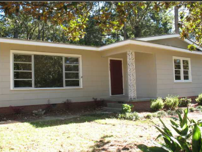 A small house with a red door and white trim is surrounded by greenery.