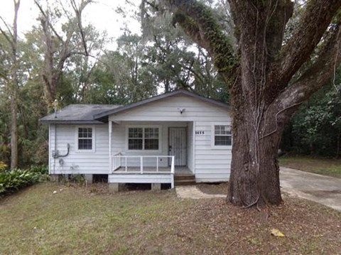 A small white house with a porch and a tree in front.