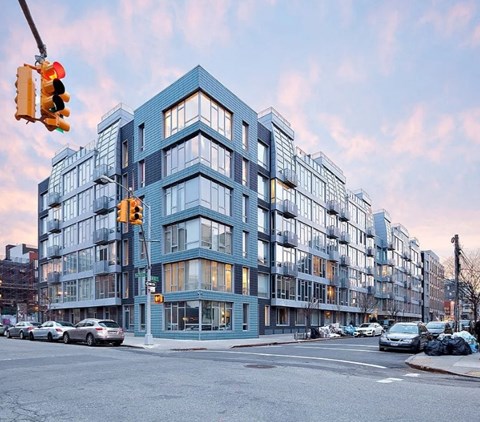 A modern multi-story building with a blue tinted facade is situated on a street corner with cars parked in front.