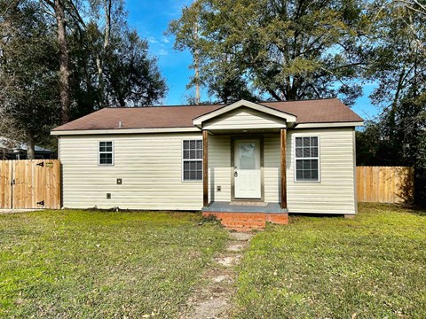 A small house with a brown roof and a white door is surrounded by a grassy area and a wooden fence.