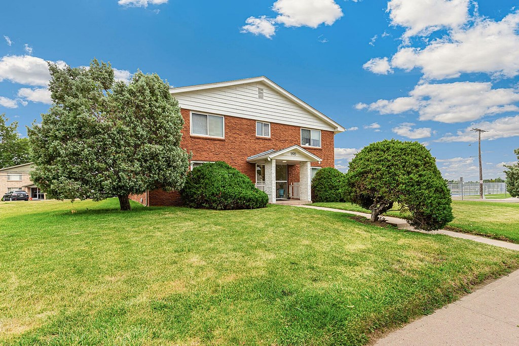 A house with a red brick exterior and a white roof is surrounded by a green lawn and trees.