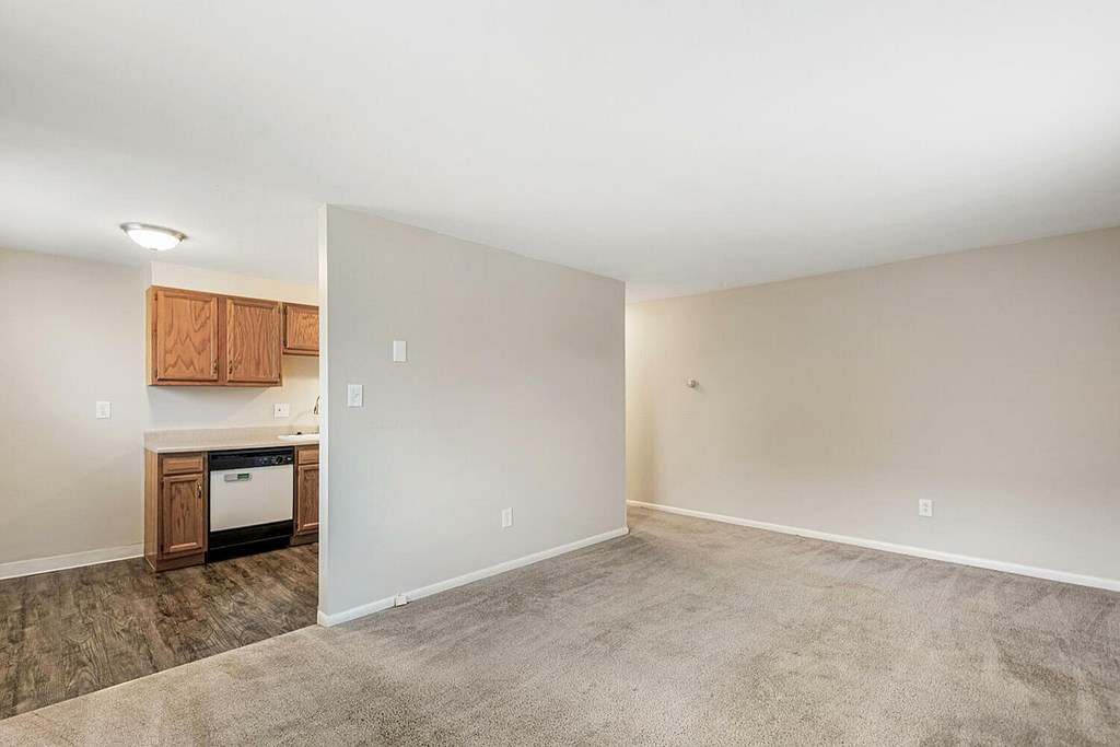 A room with a white wall and a brown floor with a small kitchen area.