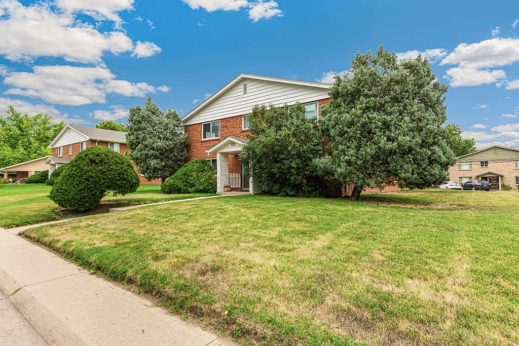 A house with a red brick exterior and a white roof is surrounded by a green lawn and trees.