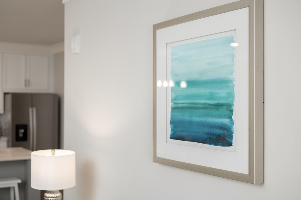 A framed artwork hangs on a wall above a table with a lit lamp.
