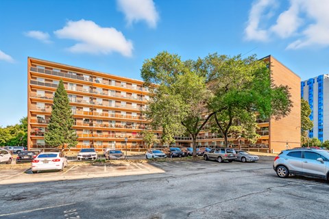A parking lot with cars and a multi-story building with trees in front.