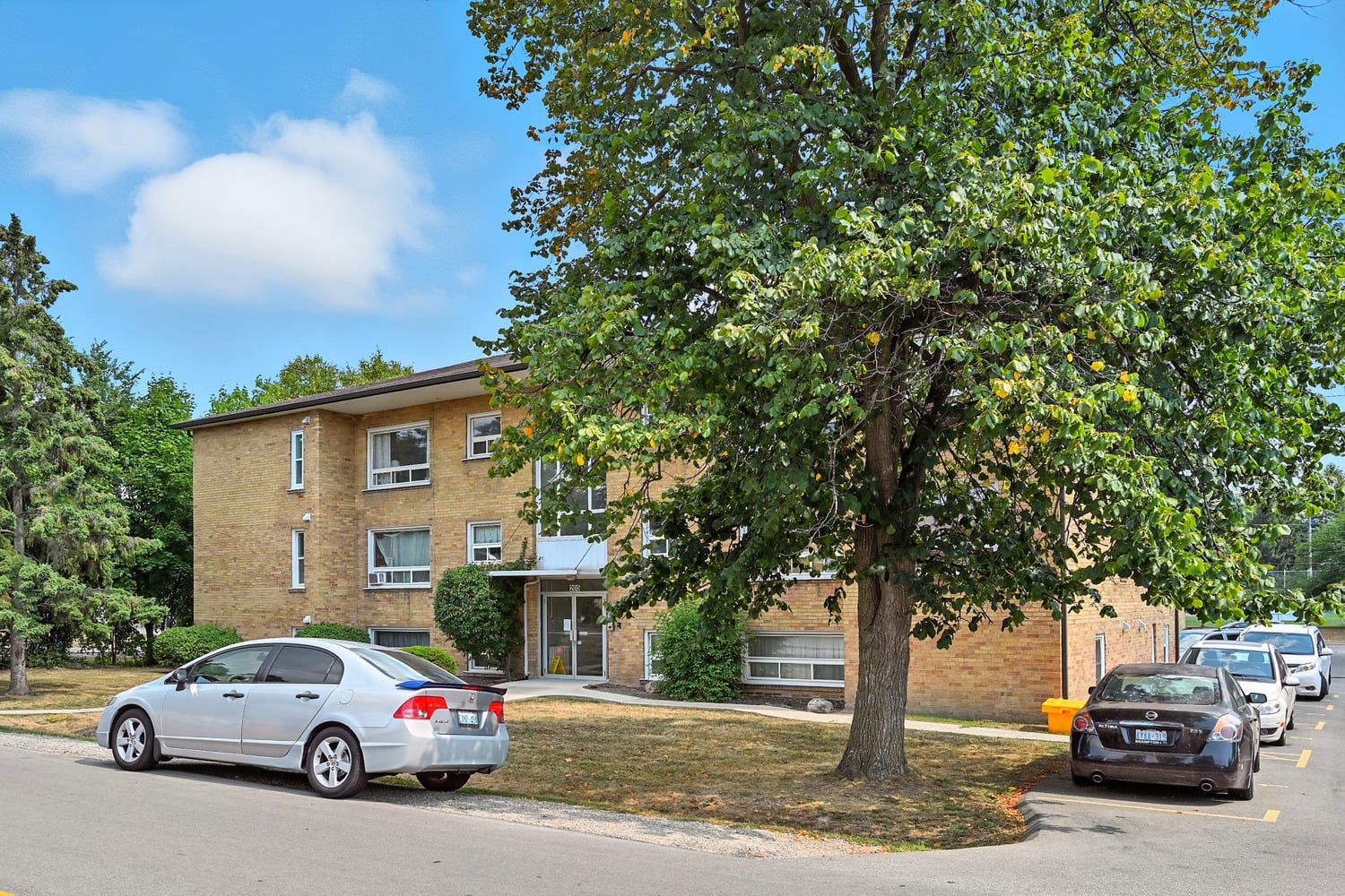 A silver car is parked on the side of the road in front of a tree and a building.