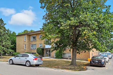 A silver car is parked on the side of the road in front of a tree and a building.