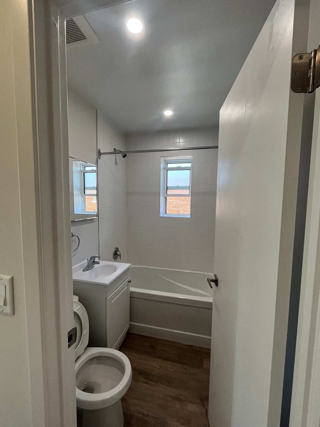 A white toilet is in a small bathroom with a white sink and a window.