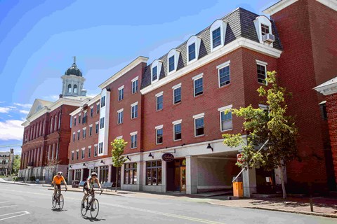 Two people riding bicycles on a street in front of a red brick building.
