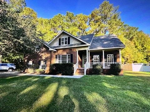 A house with a white porch and a brick exterior sits in a grassy front yard.