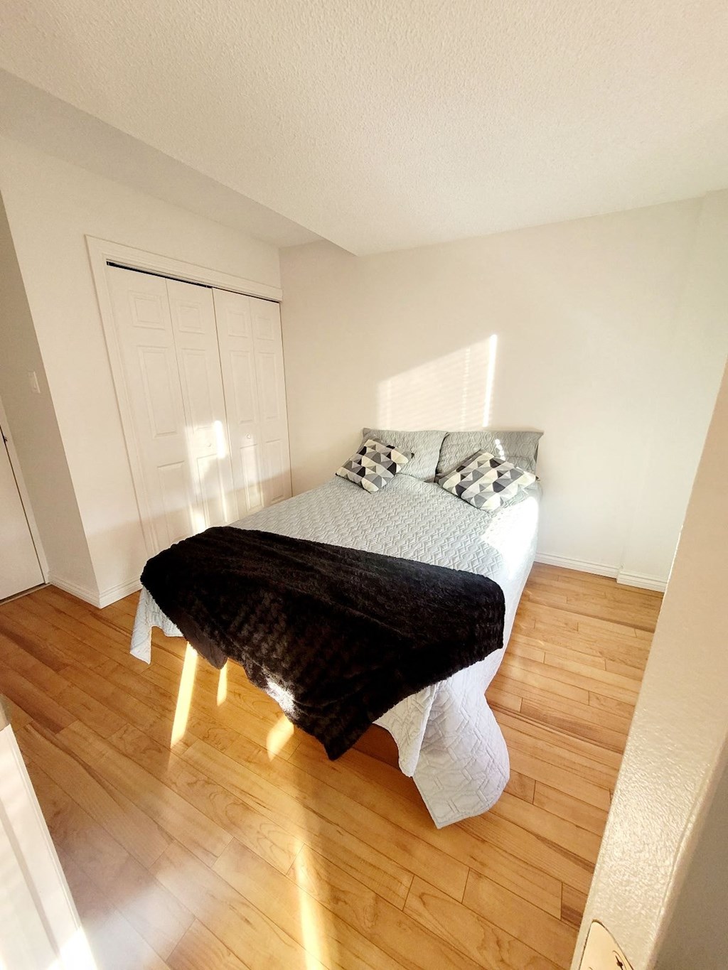A bed with a white comforter and a black blanket is in a room with wooden floors and white walls.