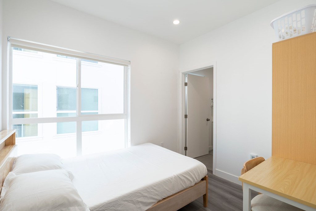 A bedroom with a bed, a desk, and a window overlooking a building.