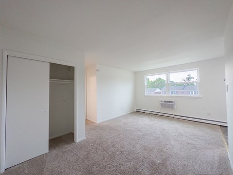 A large, empty room with a carpeted floor and a window showing a view of trees and buildings outside.