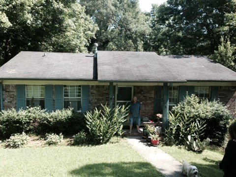A man is standing on the porch of a house with a dog lying on the ground.