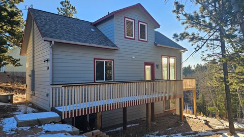 A house with a grey siding and a red roof is surrounded by trees and has a deck with a railing.