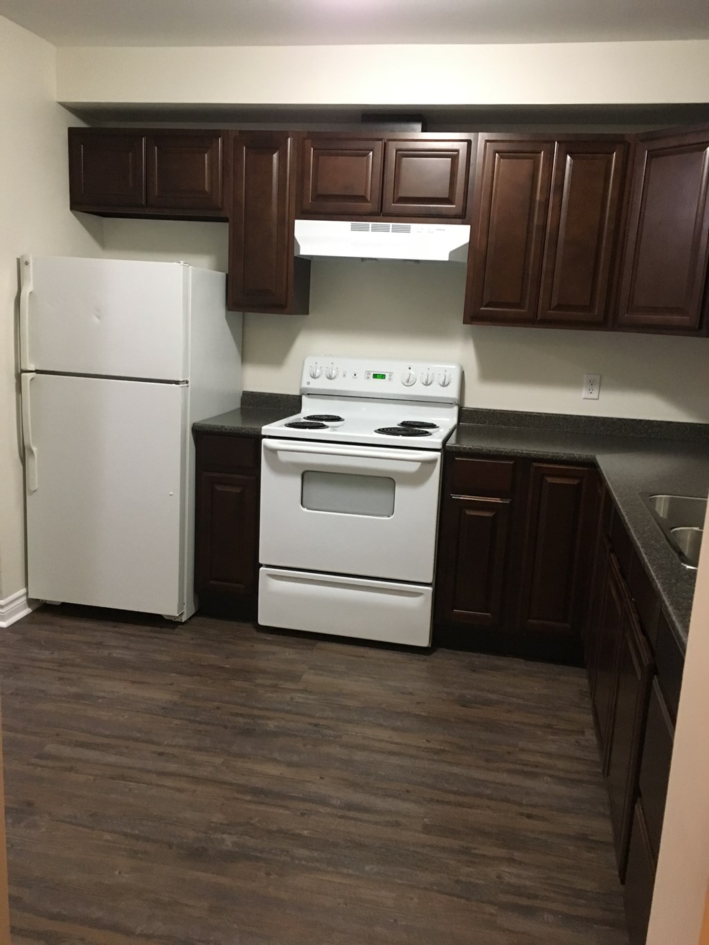 A white refrigerator stands next to a white stove in a kitchen with dark wood cabinets.