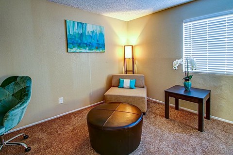 A living room with a green chair, a brown ottoman, a small couch, and a painting on the wall.