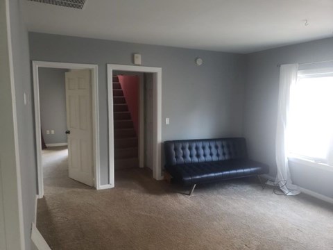 A black sofa sits in a room with a carpeted floor and a window with white curtains.
