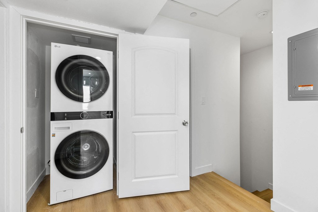 A white door is open to reveal a white dryer and washer inside a small, empty room.