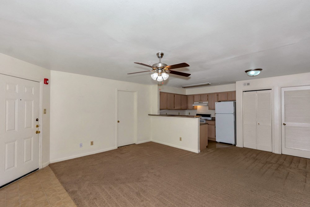 A room with a ceiling fan and a kitchenette in the background.