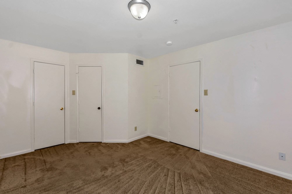 A room with white walls and brown carpeting with four doors.