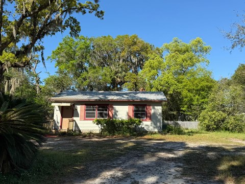 A small house with a red door and windows is surrounded by trees and vegetation.
