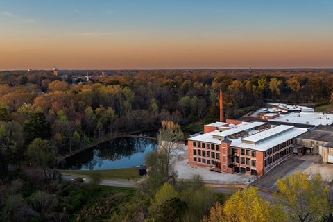 A large building with a long white chimney is surrounded by trees and a body of water.