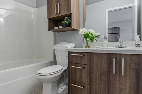 A white toilet sits in a bathroom with wooden cabinets and a white tub.