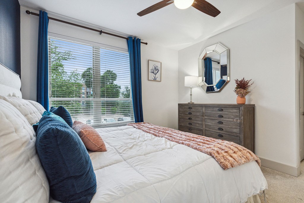 A bedroom with a bed, dresser, and window with blue curtains at Everra Midtown Park Apartments, Dallas, Texas