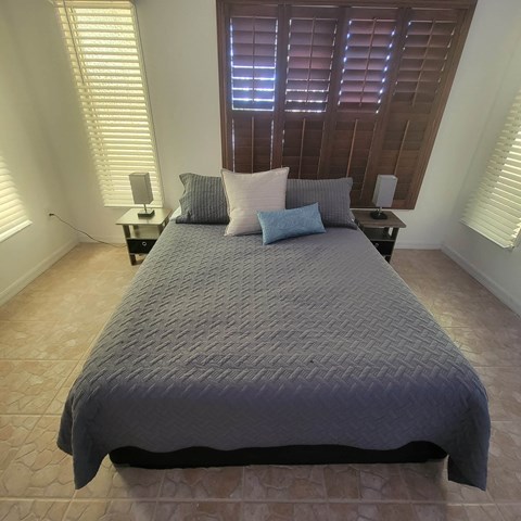 A bed with a grey comforter and two pillows sits in a room with wooden shutters on the window.