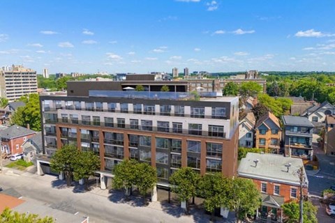 A modern multi-story building with a flat roof is surrounded by other buildings and trees.