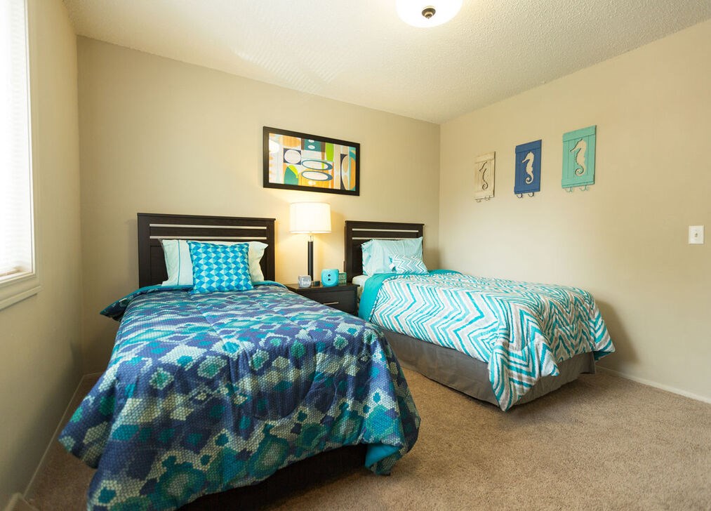 Two twin beds with blue and green bedspreads in a room with two wall art pieces.