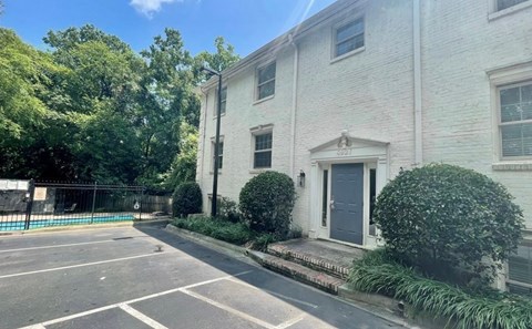 A white building with a grey door and windows is surrounded by greenery.