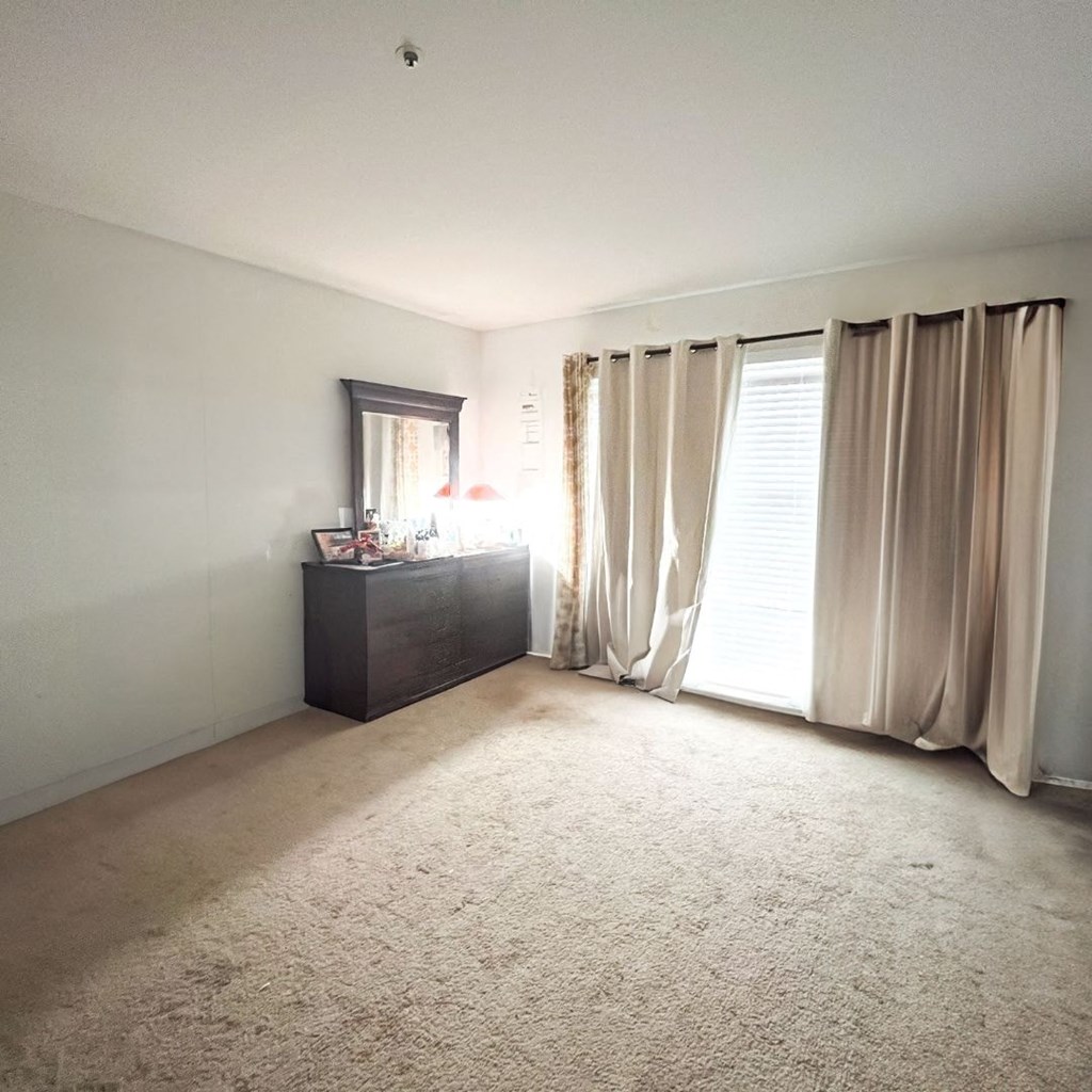 A room with a carpeted floor, a wooden cabinet, a window with curtains, and a mirror.