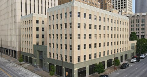 A large beige building with a green trim is situated on a busy street.