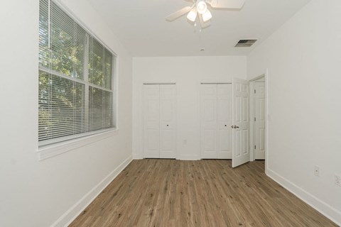 A room with white walls and wooden flooring, featuring a window with blinds, a ceiling fan, and two doors.