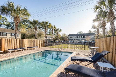 A pool surrounded by a wooden fence with a black lounge chair and a table.