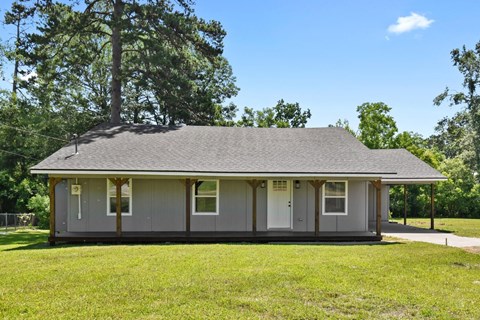 A small house with a grey roof and white trim is surrounded by green grass and trees.