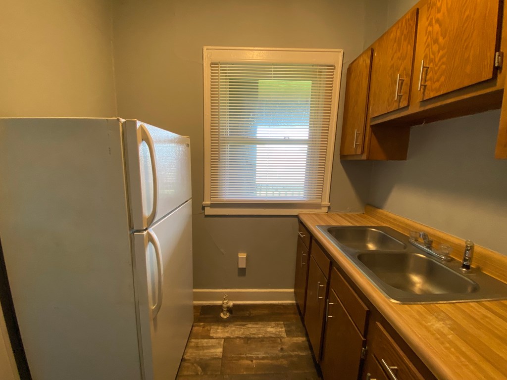 A kitchen with a white refrigerator, wooden cabinets, and a window with blinds.