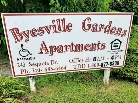 A sign for Byesville Gardens Apartments is displayed in front of some greenery.