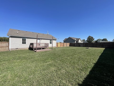 A house with a grey roof and a wooden deck is surrounded by a grassy field.