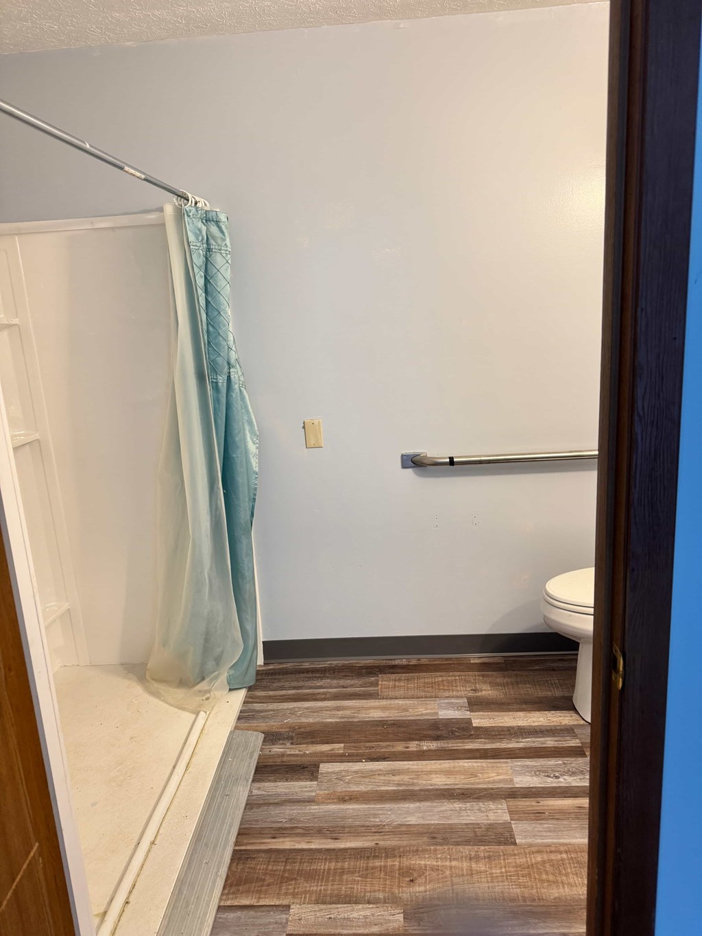 A bathroom with a toilet, a shower with a blue curtain, and a wooden floor.