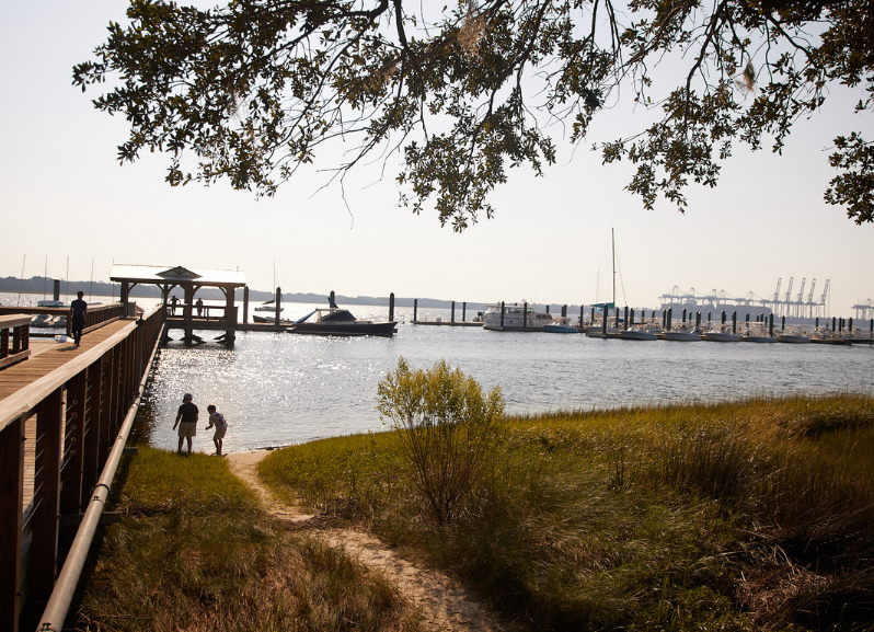 A couple walking on a boardwalk near a body of water with boats docked in the background.