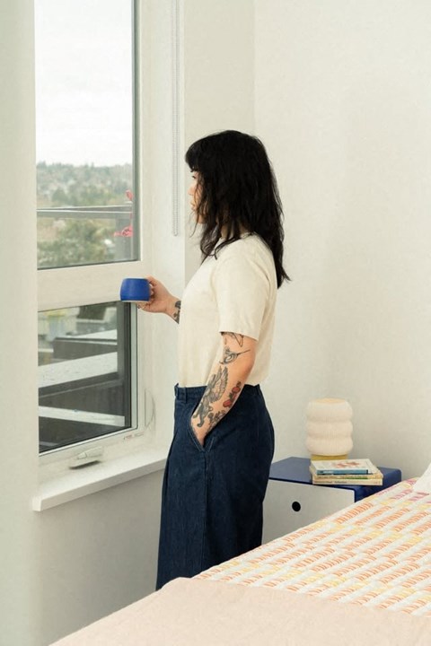 A woman with a tattoo on her arm is standing by a window holding a blue cup.