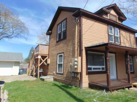 A brown house with a porch and a tree in the background.