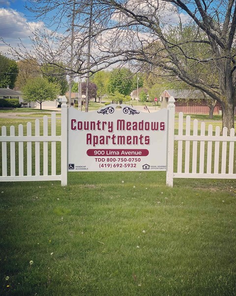 A sign for Country Meadows Apartments is displayed on a white fence.