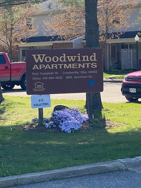 A sign for Woodwind Apartments is displayed in front of a tree.