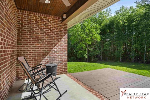 A wooden deck with a chair and a trash can is in front of a brick wall and a forest.